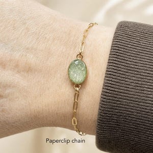 May include: A gold-toned paperclip chain bracelet featuring a central oval-shaped charm with a textured, light green, speckled surface. The charm is securely linked to the delicate metal chain, which displays a repeating elongated link design.