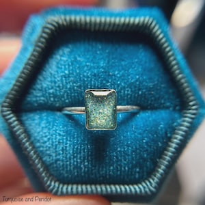 May include: A silver ring with a rectangular, turquoise-colored stone, set against a teal velvet ring box. The stone has a speckled appearance with hints of gold and green. The text "Turquoise and Peridot" is at the bottom.