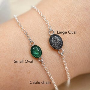 May include: Two silver cable chain bracelets featuring oval-shaped charms. One bracelet has a small oval charm with a green glitter finish, and the other has a large oval charm with a dark grey glitter finish. Text labels read Small Oval, Large Oval, and Cable chain.