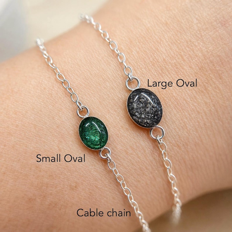 May include: Two silver cable chain bracelets featuring oval-shaped charms. One bracelet has a small oval charm with a green glitter finish, and the other has a large oval charm with a dark grey glitter finish. Text labels read Small Oval, Large Oval, and Cable chain.