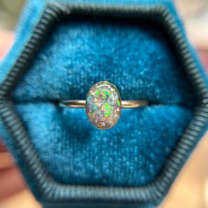 May include: A ring featuring an oval-shaped stone with a dark, translucent base and vibrant, multi-colored iridescent flecks of green, orange, and blue. The stone is set in a simple, thin, polished metallic band that encircles the finger.