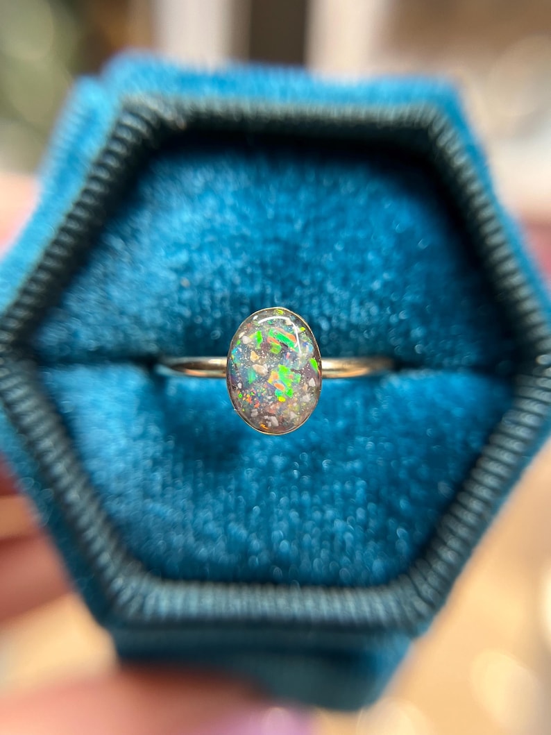 May include: A ring featuring an oval-shaped stone with a dark, translucent base and vibrant, multi-colored iridescent flecks of green, orange, and blue. The stone is set in a simple, thin, polished metallic band that encircles the finger.