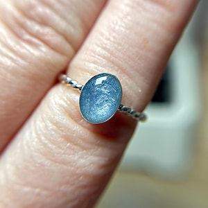 May include: A ring featuring a smooth, oval-shaped cabochon stone in a shimmering, metallic light blue color. The stone is set in a polished silver-toned metal band that has a distinct twisted, rope-like texture around the entire circumference.