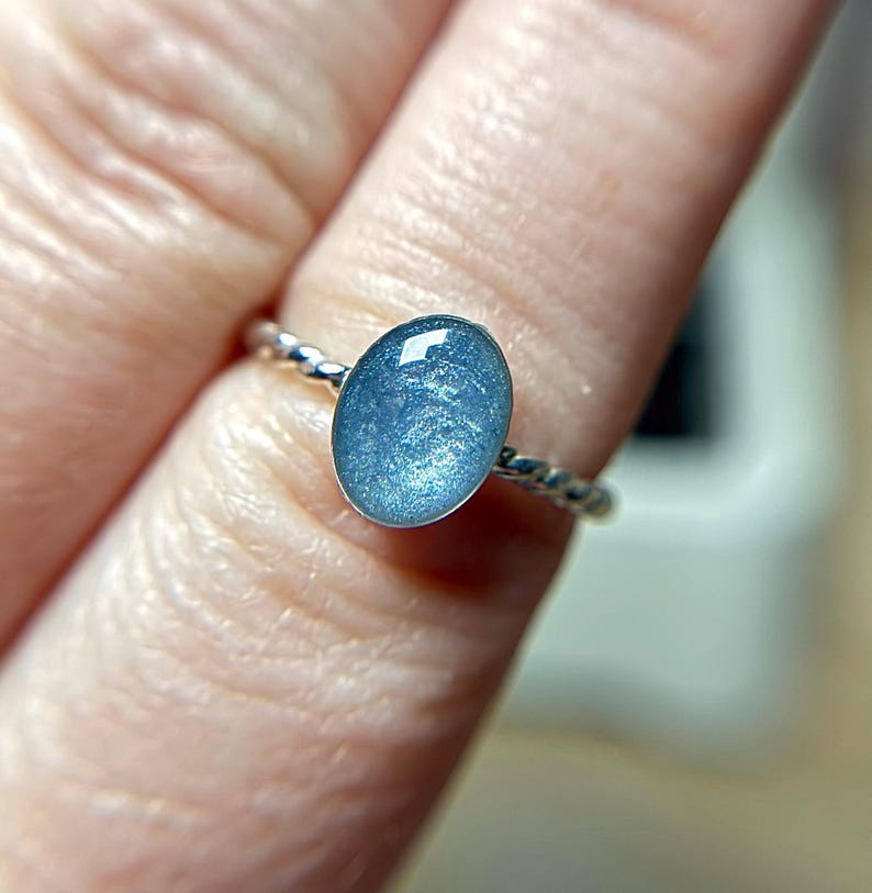 May include: A ring featuring a smooth, oval-shaped cabochon stone in a shimmering, metallic light blue color. The stone is set in a polished silver-toned metal band that has a distinct twisted, rope-like texture around the entire circumference.