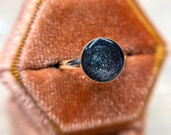 May include: A silver-toned ring featuring a circular setting with a dark, midnight blue surface embedded with fine, shimmering glitter particles that catch the light, creating a starry, celestial appearance on the face of the jewelry piece.