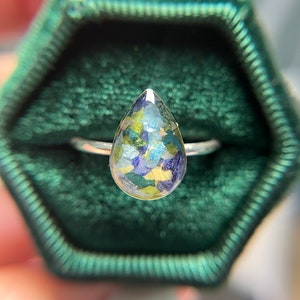 K&ouml;nnte beinhalten: Ein tropfenf&ouml;rmiger Ring mit einem silbernen Band und einem klaren Harzstein, gef&uuml;llt mit gepressten Blumen in Gr&uuml;n-, Blau- und Gelbt&ouml;nen.