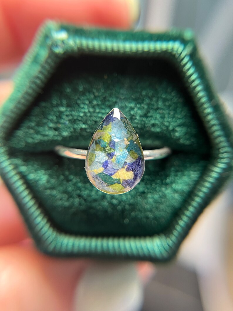 K&ouml;nnte beinhalten: Ein tropfenf&ouml;rmiger Ring mit einem silbernen Band und einem klaren Harzstein, gef&uuml;llt mit gepressten Blumen in Gr&uuml;n-, Blau- und Gelbt&ouml;nen.
