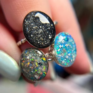 May include: Three rings featuring oval-shaped settings with textured, multi-colored inlays. One ring has a black base with light flecks, one has a dark grey base with iridescent rainbow fragments, and one has a bright blue base with silver flakes.