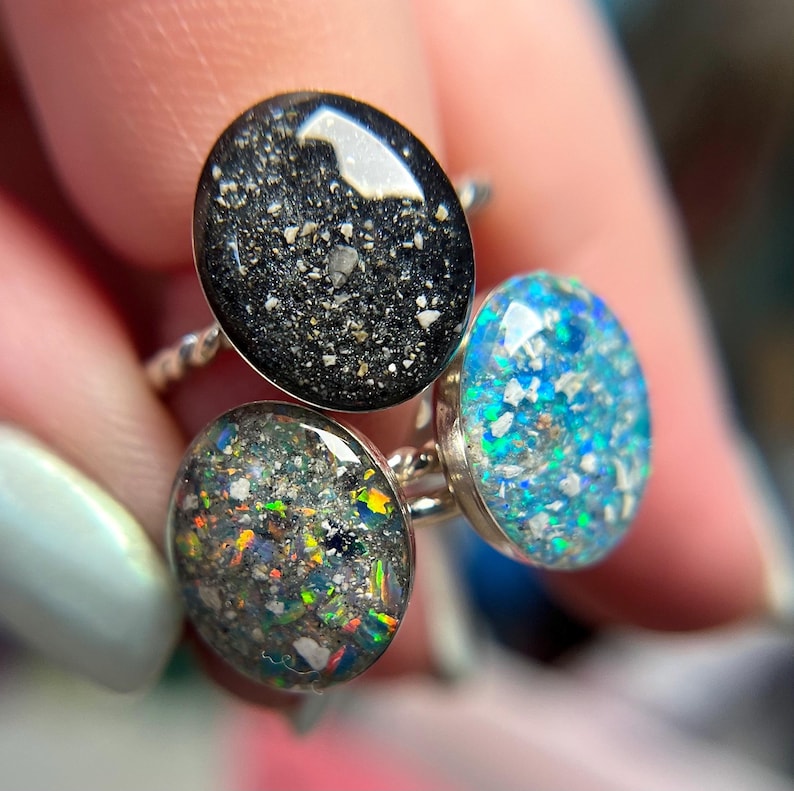 May include: Three rings featuring oval-shaped settings with textured, multi-colored inlays. One ring has a black base with light flecks, one has a dark grey base with iridescent rainbow fragments, and one has a bright blue base with silver flakes.