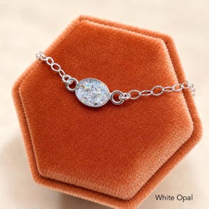 May include: A silver-toned chain bracelet featuring a central oval-shaped charm with a white opal-like finish. The charm displays a textured, iridescent surface with flecks of light-reflecting material. The text White Opal is visible.