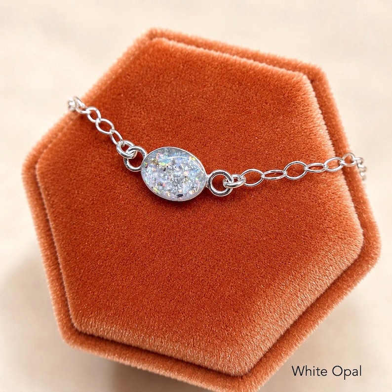 May include: A silver-toned chain bracelet featuring a central oval-shaped charm with a white opal-like finish. The charm displays a textured, iridescent surface with flecks of light-reflecting material. The text White Opal is visible.