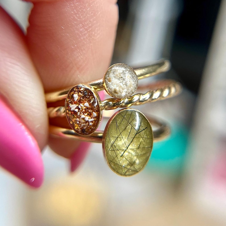 May include: A stack of three gold rings featuring distinct oval and circular settings. One ring has a light beige textured inlay, another displays a copper-toned speckled surface, and the third features a translucent olive green stone with black inclusions.