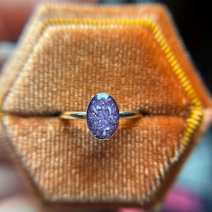 May include: A ring featuring a thin, polished metal band and an oval-shaped setting. The center stone is a deep purple color with embedded iridescent, glitter-like flecks that catch the light, creating a textured and sparkling appearance.