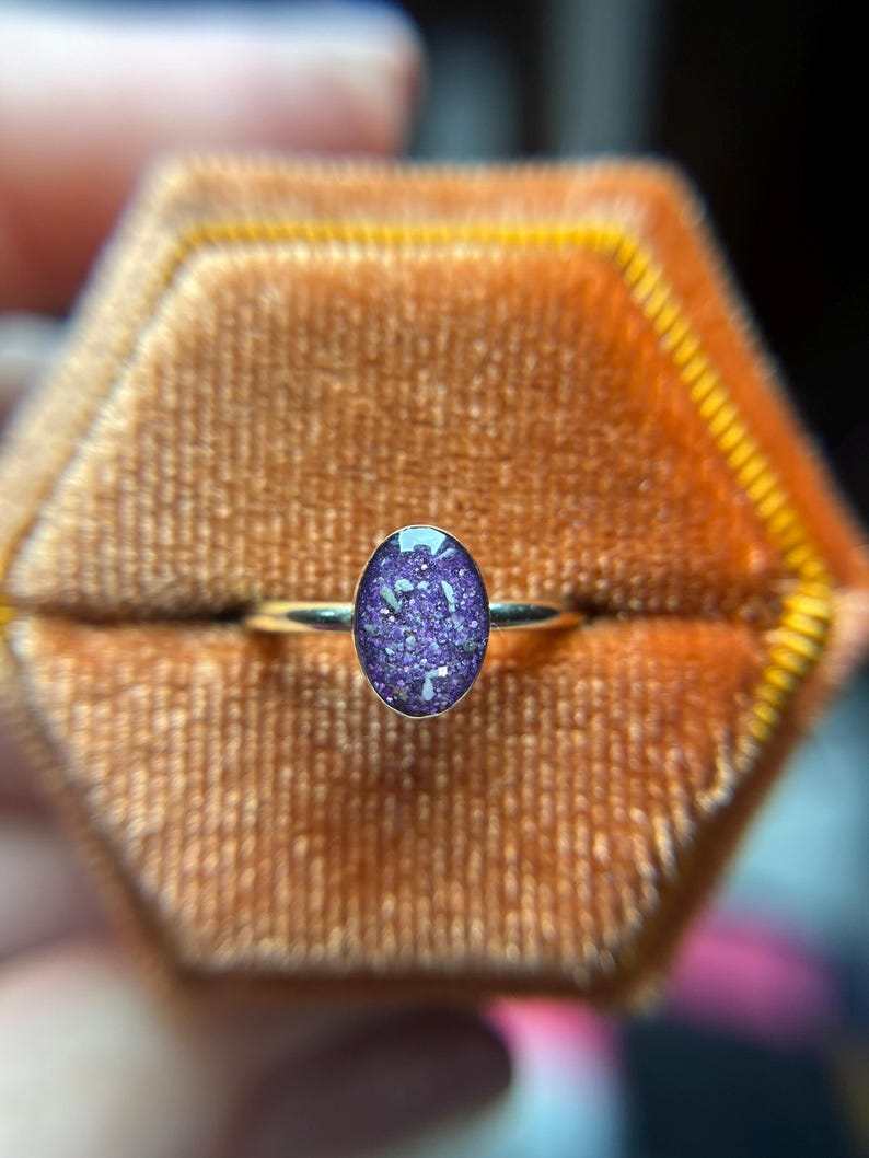 May include: A ring featuring a thin, polished metal band and an oval-shaped setting. The center stone is a deep purple color with embedded iridescent, glitter-like flecks that catch the light, creating a textured and sparkling appearance.
