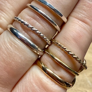 May include: A collection of six thin metal rings worn on fingers. The set includes three silver-toned rings, featuring one hammered band, one smooth band, and one twisted rope-style band, alongside three gold-toned rings with similar smooth and twisted designs.