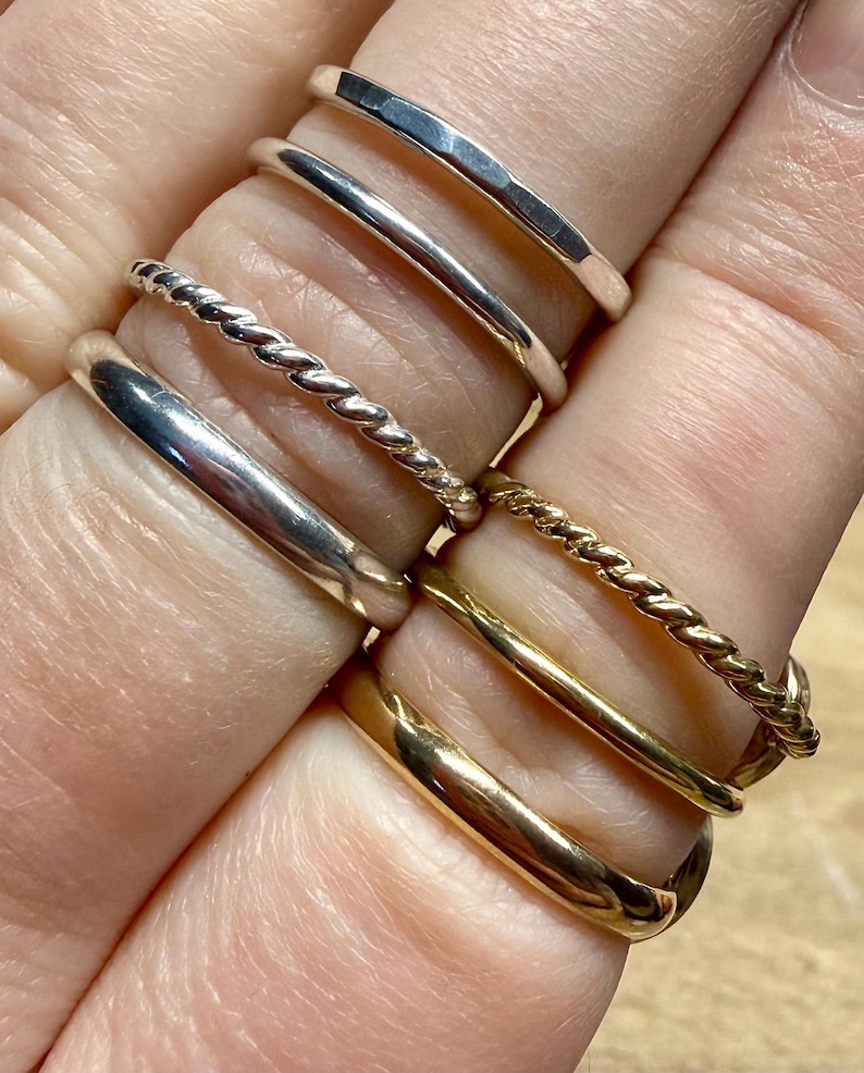 May include: A collection of six thin metal rings worn on fingers. The set includes three silver-toned rings, featuring one hammered band, one smooth band, and one twisted rope-style band, alongside three gold-toned rings with similar smooth and twisted designs.