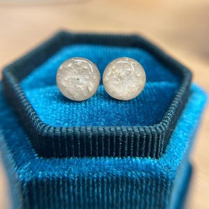 May include: A pair of round stud earrings featuring a smooth, glossy surface with a speckled, off-white and beige textured appearance. The circular earrings have a polished finish and are presented side by side, showcasing a neutral color palette.