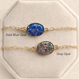May include: Two delicate gold-tone chain bracelets featuring oval-shaped pendants. The top bracelet has a dark blue opal inlay, and the bottom bracelet has a gray opal inlay. The text "Dark Blue Opal" and "Gray Opal" is printed near each piece.