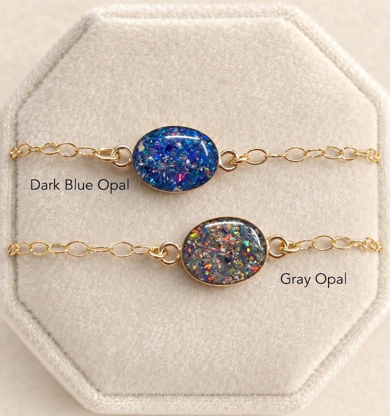 May include: Two delicate gold-tone chain bracelets featuring oval-shaped pendants. The top bracelet has a dark blue opal inlay, and the bottom bracelet has a gray opal inlay. The text "Dark Blue Opal" and "Gray Opal" is printed near each piece.