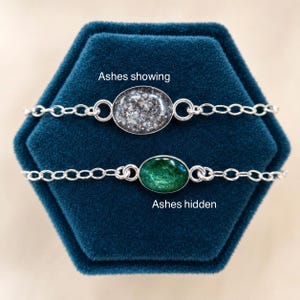 May include: Two silver chain bracelets with oval pendants. The top pendant has a textured gray and white surface labeled 'Ashes showing'. The bottom pendant has a smooth, glossy green surface labeled 'Ashes hidden'. Both feature delicate metal links.