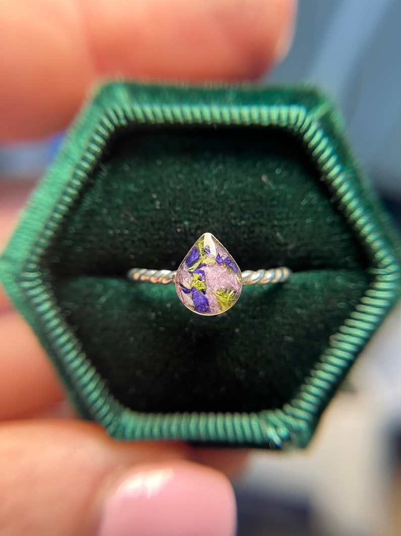 K&ouml;nnte beinhalten: Ein silberner Ring mit einer tropfenf&ouml;rmigen Harzfassung, gef&uuml;llt mit gepressten lila, rosa und gr&uuml;nen Blumen.