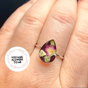 K&ouml;nnte beinhalten: Ein tropfenf&ouml;rmiger Ring mit einem silbernen Band und einer klaren Harzfassung, die gepresste Blumen in Rosa-, Gelb- und Gr&uuml;nt&ouml;nen enth&auml;lt.