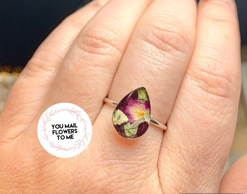 K&ouml;nnte beinhalten: Ein tropfenf&ouml;rmiger Ring mit einem silbernen Band und einer klaren Harzfassung, die gepresste Blumen in Rosa-, Gelb- und Gr&uuml;nt&ouml;nen enth&auml;lt.
