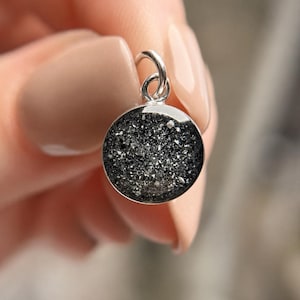 May include: A circular pendant featuring a textured onyx-colored inlay with visible flecks, set in a silver-toned metal frame with a jump ring. The pendant measures 8mm in diameter and includes the text 8mm Ashes Showing Color: Onyx.