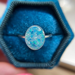 May include: A silver-toned ring featuring an oval-shaped setting filled with a mosaic of iridescent blue, light blue, and white flecks. The smooth, rounded surface of the inlay creates a reflective finish, set against a thin, polished metal band.