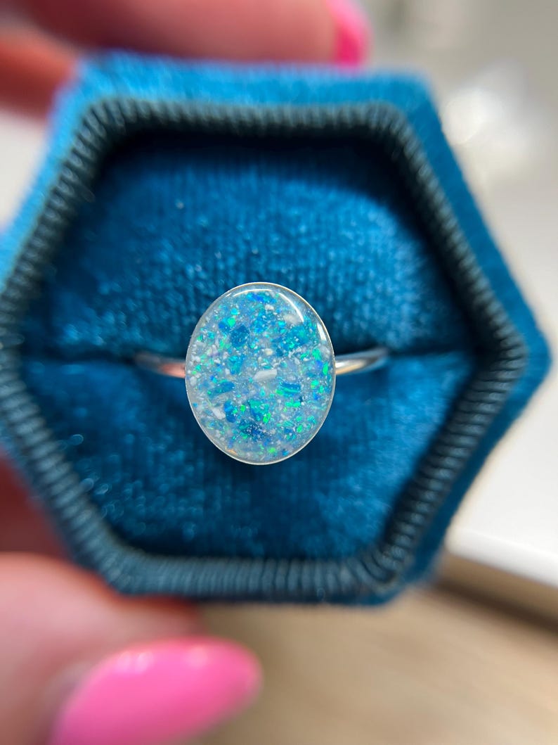 May include: A silver-toned ring featuring an oval-shaped setting filled with a mosaic of iridescent blue, light blue, and white flecks. The smooth, rounded surface of the inlay creates a reflective finish, set against a thin, polished metal band.