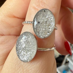 May include: Two silver-toned rings featuring oval-shaped settings filled with a textured, shimmering grey and silver glitter material. One ring has a twisted metal band, while the other ring features a smooth, plain polished silver-toned metal band.