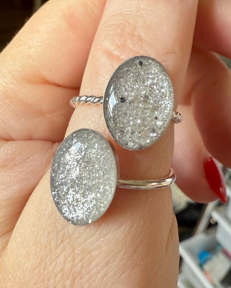 May include: Two silver-toned rings featuring oval-shaped settings filled with a textured, shimmering grey and silver glitter material. One ring has a twisted metal band, while the other ring features a smooth, plain polished silver-toned metal band.