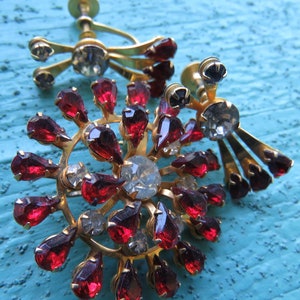 May include: A pair of vintage earrings with a gold tone metal base and red and clear rhinestones. The earrings are in the shape of a starburst with a cluster of red rhinestones in the center.