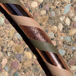 Pu&ograve; includere: Un hula hoop con una base in rame metallizzato avvolto da strisce diagonali di nastro verde oliva, beige e nastro glitterato color rame scintillante. L'oggetto circolare mostra un motivo ripetitivo di queste tre texture e colori.