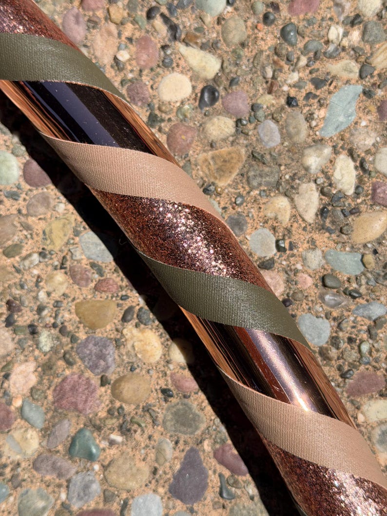 Pu&ograve; includere: Un hula hoop con una base in rame metallizzato avvolto da strisce diagonali di nastro verde oliva, beige e nastro glitterato color rame scintillante. L'oggetto circolare mostra un motivo ripetitivo di queste tre texture e colori.