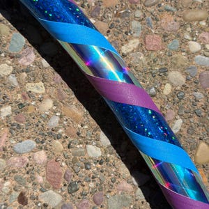 Pu&ograve; includere: Un hula hoop circolare caratterizzato da una base blu iridescente riflettente con una finitura glitter olografica. Il cerchio &egrave; avvolto in un motivo a spirale con strisce alternate di nastro viola opaco e nastro in tessuto blu ciano.