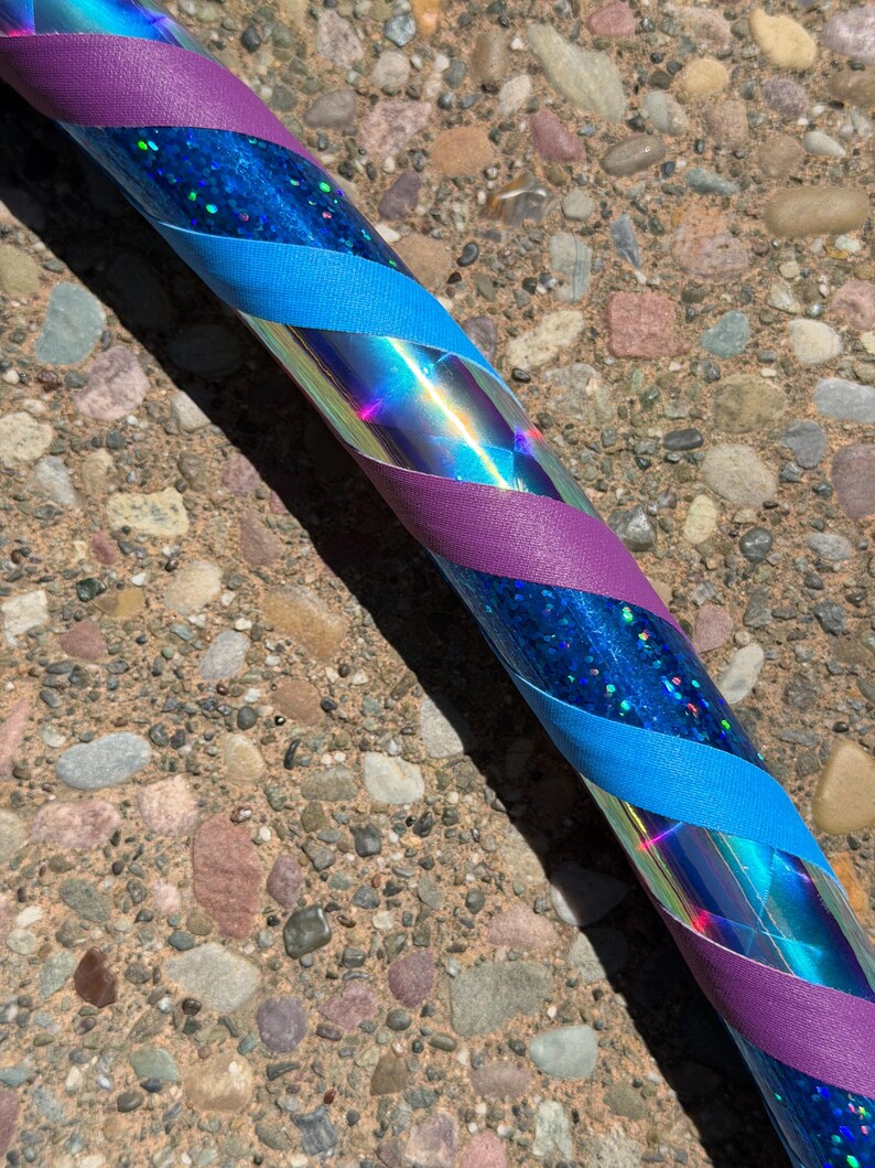 Pu&ograve; includere: Un hula hoop circolare caratterizzato da una base blu iridescente riflettente con una finitura glitter olografica. Il cerchio &egrave; avvolto in un motivo a spirale con strisce alternate di nastro viola opaco e nastro in tessuto blu ciano.