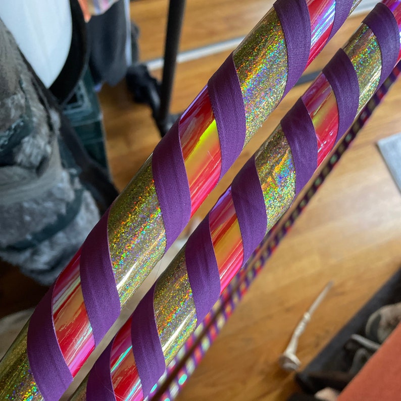 Pu&ograve; includere: Due hula hoop caratterizzati da una finitura olografica iridescente rossa e dorata. Ogni cerchio &egrave; avvolto da un nastro in tessuto viola opaco a spirale, che crea un netto contrasto con la superficie metallica scintillante e multicolore.