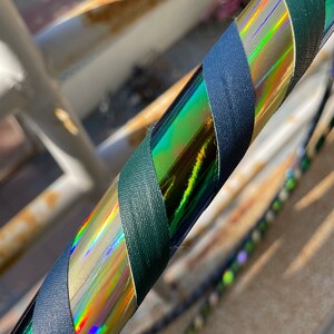 Pu&ograve; includere: Un hula hoop circolare caratterizzato da un design a spirale con nastro testurizzato verde scuro che si alterna a un nastro olografico iridescente e riflettente, che mostra uno spettro di colori quando la luce colpisce la superficie.