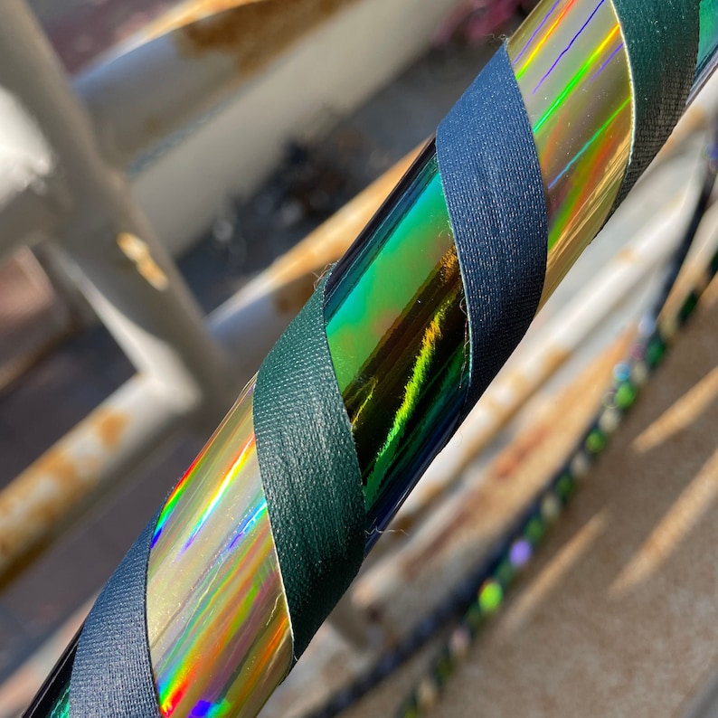 Pu&ograve; includere: Un hula hoop circolare caratterizzato da un design a spirale con nastro testurizzato verde scuro che si alterna a un nastro olografico iridescente e riflettente, che mostra uno spettro di colori quando la luce colpisce la superficie.