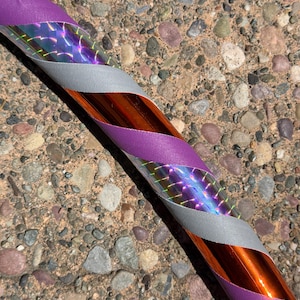 Pu&ograve; includere: Un hula hoop con una base arancione metallizzata avvolta in un motivo a spirale di nastro in tessuto viola, nastro in tessuto argentato e una pellicola olografica che riflette la luce con un effetto prismatico sulla superficie del cerchio.