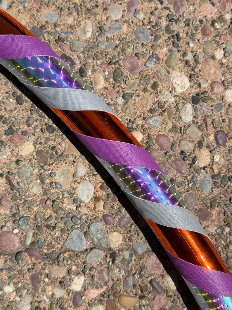 Pu&ograve; includere: Un hula hoop con una base arancione metallizzata avvolta in un motivo a spirale di nastro in tessuto viola, nastro in tessuto argentato e una pellicola olografica che riflette la luce con un effetto prismatico sulla superficie del cerchio.