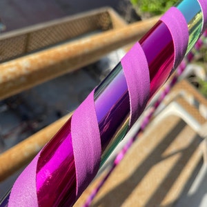 Pu&ograve; includere: Un hula hoop caratterizzato da una superficie metallica riflettente e iridescente color viola e verde acqua, avvolto in un motivo a spirale con un nastro in tessuto viola opaco e tinta unita. Il tubo circolare presenta una finitura liscia.