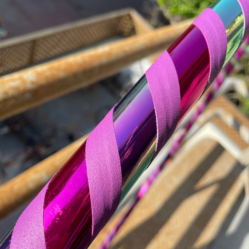 Pu&ograve; includere: Un hula hoop caratterizzato da una superficie metallica riflettente e iridescente color viola e verde acqua, avvolto in un motivo a spirale con un nastro in tessuto viola opaco e tinta unita. Il tubo circolare presenta una finitura liscia.