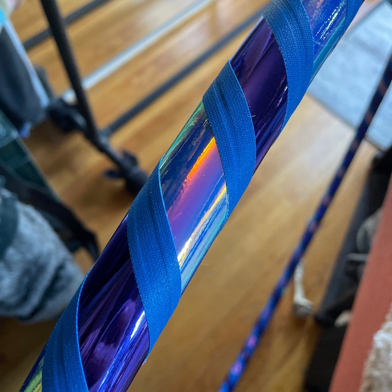 Pu&ograve; includere: Un hula hoop circolare caratterizzato da una superficie viola iridescente e riflettente che cambia colore. Il cerchio &egrave; avvolto da un nastro in tessuto blu, solido e testurizzato, applicato con un motivo a spirale diagonale lungo il tubo.
