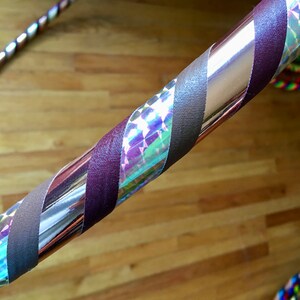 Pu&ograve; includere: Un hula hoop circolare caratterizzato da un design a spirale con nastro adesivo marrone scuro opaco e nastro olografico argentato riflettente. La superficie dell'hula hoop alterna strisce marrone scuro a sezioni argentate prismatiche.