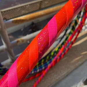 Pu&ograve; includere: Un hula hoop con un motivo a spirale di bande alternate. Il design &egrave; composto da nastro glitterato arancione brillante, nastro glitterato rosa vivace e nastro rosa opaco a tinta unita avvolti diagonalmente attorno alla struttura tubolare.