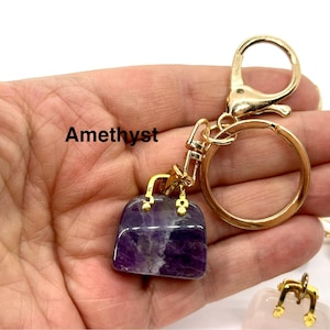 May include: Amethyst gemstone handbag keychain with a gold-tone metal chain and ring. The keychain is held in a hand.