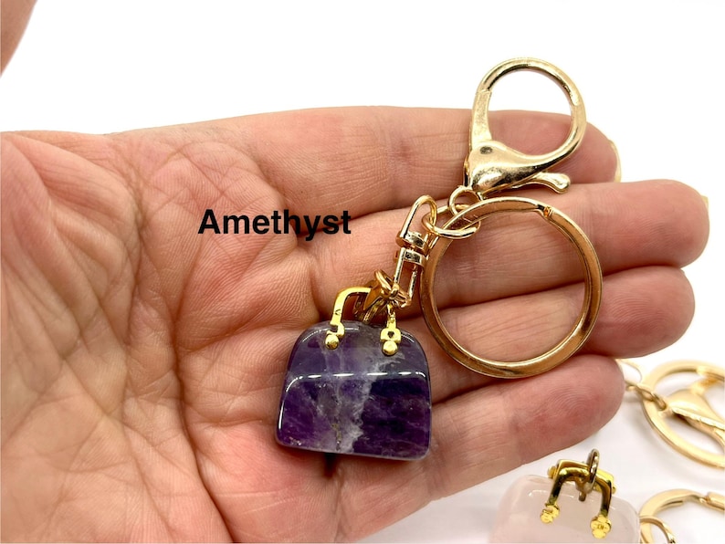May include: Amethyst gemstone handbag keychain with a gold-tone metal chain and ring. The keychain is held in a hand.
