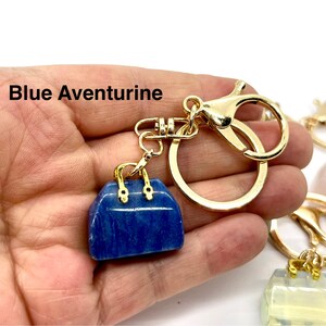 May include: A gold-tone keychain with a blue aventurine handbag charm. The charm is a small, detailed replica of a handbag with gold-tone accents. The keychain is attached to a gold-tone split ring.